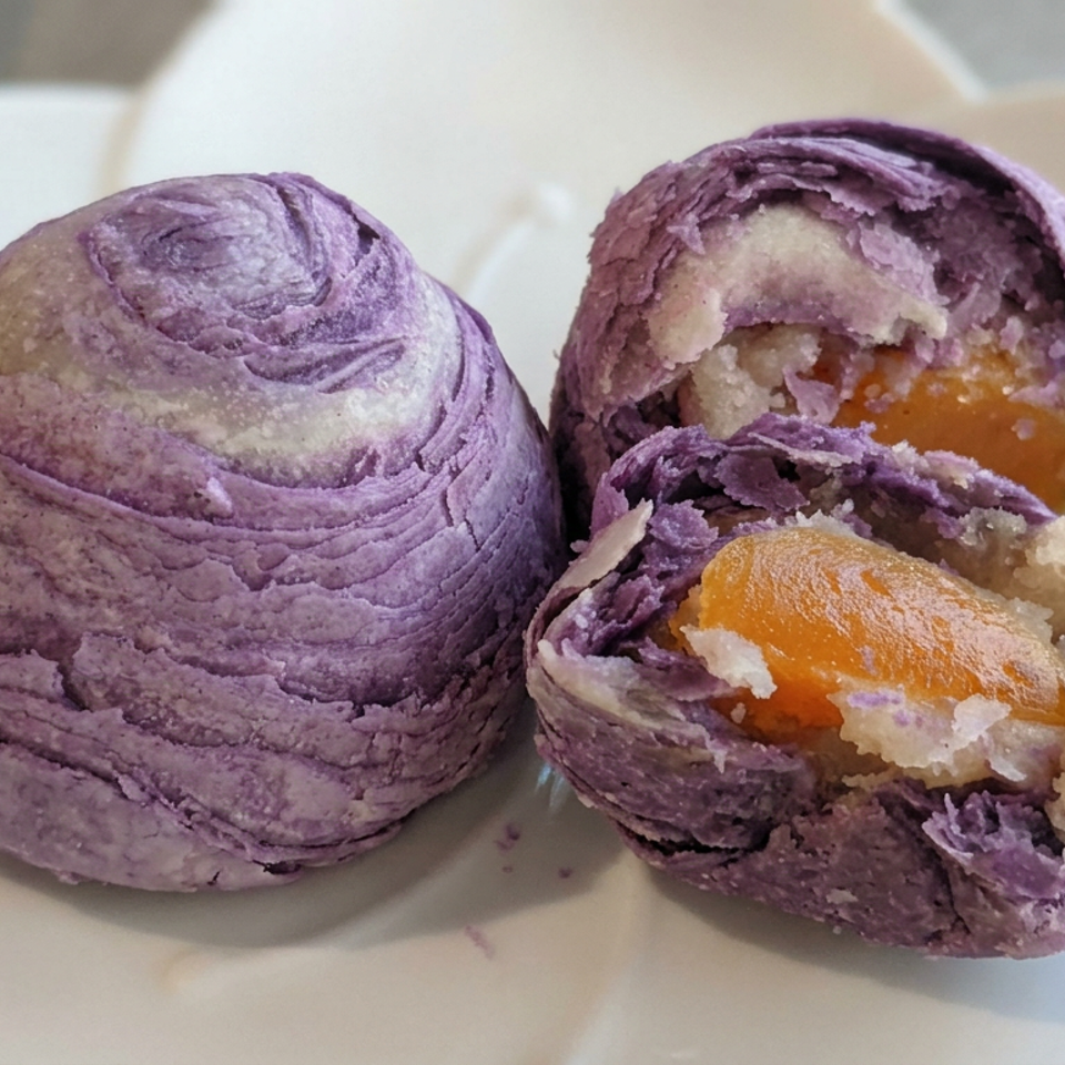 Two purple layered pastries sit on a white plate, one cut open to reveal orange fruit filling and crumbly interior.